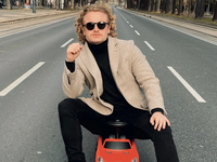 Der österreichischer Kabarettist Paulus Bohl trägt eine schwarze Sonnenbrille und sitzt auf einem roten Spielzeugauto.